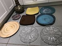 Seven serving dishes arranged on floor including one wooden plate, three pottery plates (blue-green, dark brown square, yellow square), and three clear cut-glass plates.