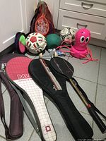 Photo showing three tennis rackets with covers placed on grey tiled floor, along with balls and other sports equipment in background.