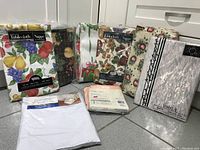 Photo of eight vinyl tablecovers showing different patterns including fruits, florals, and abstract designs. Most are in packaging with label visible.