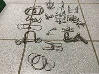 Overview of collection of 14 different metal wire brain teaser puzzles arranged on tiled floor, showing variety of shapes and sizes.
