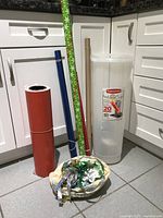 Wide shot showing four rolls of wrapping paper standing upright and leaning in a corner, a bowl with assorted ribbons in front, and Rubbermaid container in background.