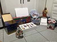 Photo showing multiple Christmas decorative items including snow globes, figurines, unopened boxes of ornaments, and a Jesus figurine statue arranged on the floor.