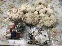 Multiple balls of white and off-white yarn along with spools of sewing thread and bags of buttons on floral background