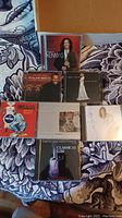 Photo shows a collection of 8 CDs featuring artists like Kenny G, Carly Simon, and a Motown tribute album on a patterned fabric surface.