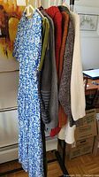 Full view of six clothing items hanging on a rack including a blue and white floral dress and five sweaters in various colors and textures.