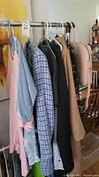 Clothing items hanging on a rack including pink and grey tops, blue and white tweed jacket, black blazer, tan coat, and light blue jeans.