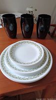 Corelle dishes and black vases displayed on wooden table. The dishes are white with green leaf pattern on the rims. Three tall black glossy vases are placed behind the dishes.