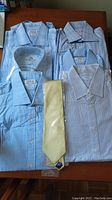Six men's dress shirts folded and laid flat on a wooden surface with one yellow tie placed vertically in the center.