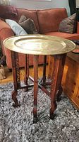Full view of the folding table showing brass tray top resting on wooden foldable base with carved decorative elements.