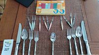 Full display of two sets of Japanese flatware with knives, spoons, forks, small spoon, pickle forks, and vintage beverage coasters on woven mat surface.