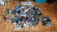 Wide view of mixed Lego pieces and models spread on parquet floor with some small vehicles and airplane parts visible.