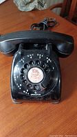 Photo showing full view of black rotary phone with handset and cord on wooden surface.