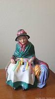 Front view of Royal Doulton figurine showing seated elderly woman stitching ribbons on white cloth, colorful ribbons draped by her side.
