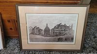 Framed black and white etching of a European town square showing multiple traditional buildings with distinctive architectural elements, displayed on carpet.