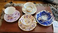 Four teacups and matching saucers shown on a wooden table, each pair with distinct floral and gold designs including purple, blue, and gold accents.