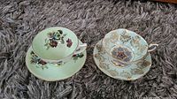Two teacups with their matching saucers placed on a carpet, one set green with floral pattern, the other pale blue with gold and ornate detailing.