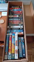 Top view of box showing 24 vintage VHS tapes in their original cases with readable titles.