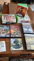 Photo of stacked Xbox One and Xbox 360 game cases on table with some PS2 games laid out alongside, showing titles and condition of cases.