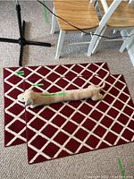 Two red and white criss-cross patterned floor rugs placed on carpeted floor with beige dachshund-shaped dog door draft protector on top.
