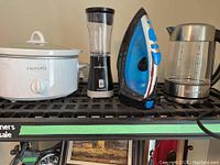 Photo showing four appliances: white Crockpot, black and clear Hamilton Beach blender, blue and white Sunbeam iron, metallic and clear Thinkkitchen kettle arranged on shelf.