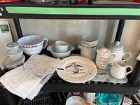 Various kitchenware items including bowls, plates, teapot, creamers, canisters, tablecloths, and a cutting board arranged on a black metal rack.