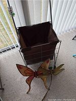 Full view of metal shopping buggy with brown fabric liner and two metal wall decor pieces in front, one butterfly and one bird shaped.