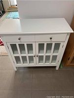 Front view of the white cabinet showing the drawer at the top and two glass paneled doors below.