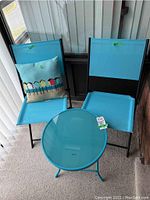 Two blue folding chairs with black metal frames, round blue table with tempered glass top, and decorative pillow arranged on balcony.