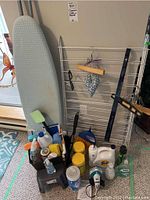 Full view of the ironing board with blue checkered cover, a folding metal drying rack, and a basket holding assorted cleaning supplies and an iron on the floor.