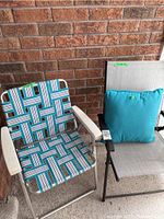 Two folding patio chairs and one blue pillow photographed on balcony against a brick wall. One chair has blue/white woven webbing, the other is grey mesh with armrests and pillow on seat.