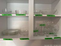Kitchen cabinet shelf with clearly labeled glass plates, bowls, and glasses stacked and grouped with green labels identifying quantities and types of dishware.