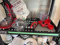 Photo of kitchen shelving with glass and plastic measuring cups, glass bowls, red kitchen utensils, and metal cooling racks.