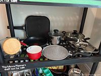 Full view of the shelf showing frying pans, pots, and glass jar with wooden lid.