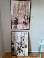 Two metal framed prints leaning against wall: top is urban couple scene in dark frame, bottom is street scene with person in red coat in lighter frame.