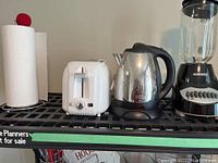 Four small kitchen appliances on a black wire shelf: paper towel holder with white paper towel, white Sunbeam 2-slice toaster, stainless steel Sunbeam electric kettle, and Black & Decker blender with glass jar and black base.