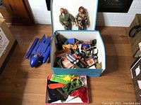 Two GI Joe action figures displayed above an open box filled with assorted plastic accessories and clothing, with a blue plastic jet vehicle beside the box on the floor.