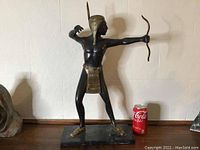 Full view of bronze statue showing Indigenous archer in stance with bow, adjacent to soda can for scale illustration.