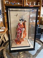 Porcelain Asian doll dressed in red and orange kimono inside a black-framed plexiglass display case with shelving and background visible.