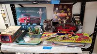 Photo shows assorted automotive collectibles on shelves including Mustang placard, Millennium coin set, diecast cars, ceramic decanter, vintage book, race car placards, John Deere collectibles.