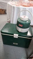 Green Coleman cooler chest with white latch on front, placed on floor next to table with glassware, showing the full chest and the beverage jug on top.