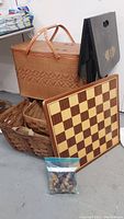 Photo showing two woven picnic baskets, one large with handles and lid, the other smaller and open weave; wooden chessboard leaning against baskets with chess pieces in bag on floor in front; black folding tray table behind them.