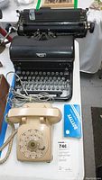 Photo showing vintage Royal manual typewriter with black body and round keys, alongside beige rotary dial telephone with handset and coiled cord on white surface.