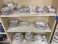 Display of entire 89 piece Sango China set on shelves showing plates, bowls, teapot, cups, saucers, creamers, sugar bowls, and platters all with pink rose pattern.