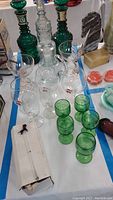 Full collection displayed on white cloth with blue tape edges including green cordial glasses, clear snifters, pink margarita glasses, water pitcher with drinking glass, especially showing the swizzle sticks in the opened box.