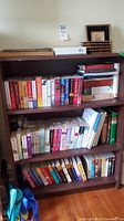 Bookshelf with multiple rows of paperback novels and cookbooks, books arranged double deep on shelves.