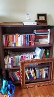 Full front view of dark brown wooden bookshelf mostly empty, showing contents excluded from sale, a variety of books in different genres and a picture frame leaning on it