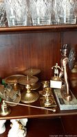 Photo showing three brass candle holders with spikes, two brass votive holders, Leonidas figurine, trophy, and snifter glass on a wooden shelf.