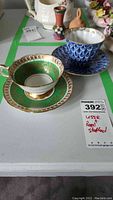 Blue and gold USSR Lomonosov porcelain tea cup and saucer alongside green and gold Royal Stafford tea cup and saucer on a white surface.