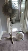 Photo showing a white floor-standing pedestal fan next to a small white table fan on a carpeted floor near a tiled bathroom.