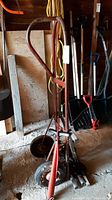 Photo of red metal hand truck with curved handle and pneumatic wheels, stored in a garage with garden tools in the background.